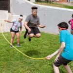 Grupo diverso de adolescentes brinca de pular corda ao ar livre em um parque ensolarado.