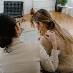Duas mulheres sentadas juntas em ambiente interno, demonstrando apoio e empatia durante uma conversa.
